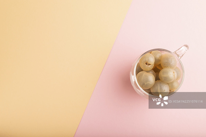 玻璃杯中成熟去皮的龙眼，置于粉橙色柔和纸张背景上，俯视，平铺，留有复制空间，极简风格图片素材