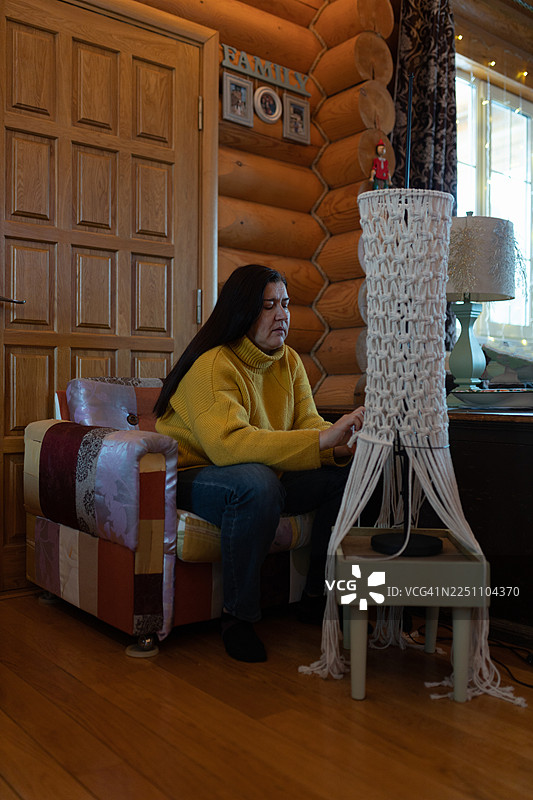 一位穿着黄色毛衣的女士坐在色彩缤纷的拼布沙发上，在舒适的木屋室内编织着毛线。图片素材