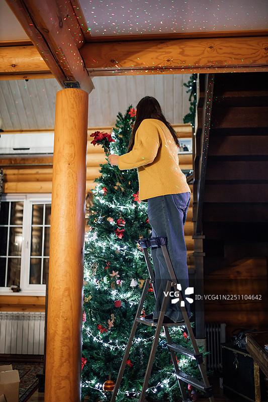 一位女士在温馨的木屋里，在节日期间爬上梯子装饰圣诞树。图片素材
