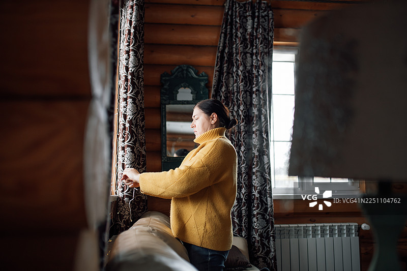 一位身穿芥末色毛衣的女士正在明亮的窗边，在舒适的小屋里调整窗帘图片素材