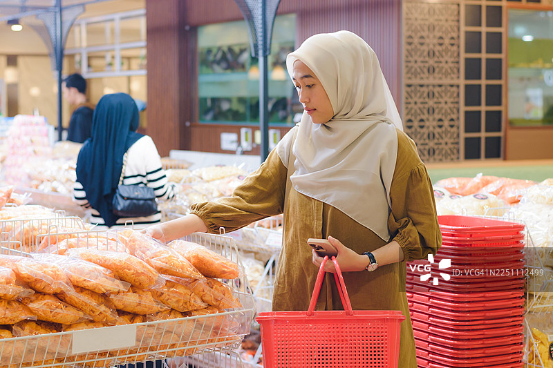 戴着头巾的穆斯林女性提着购物篮在货架上挑选零食图片素材