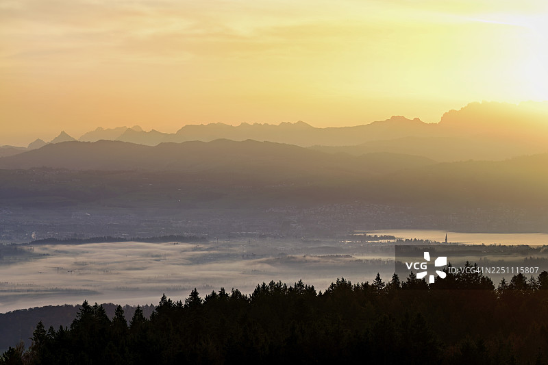 从霍本眺望楚格湖，楚格市和楚格市被大雾笼罩，其后是覆盖着积雪的弗吕布里格山和弗雷内利斯加特利山，沐浴在初升的阳光中，位于瑞士阿尔高州，拜因维尔-弗莱阿姆特。图片素材
