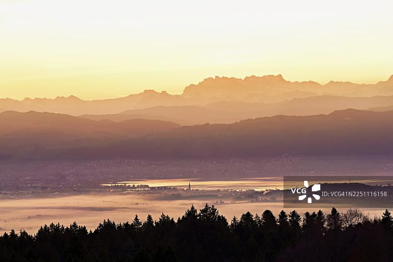 从霍本远眺楚格湖，湖畔的楚姆市和楚格市笼罩在雾气中，背后是初升太阳映照下白雪覆盖的弗吕布里格山和弗伦里斯格特利山，位于瑞士阿尔高州拜因维尔-弗莱阿姆特。图片素材