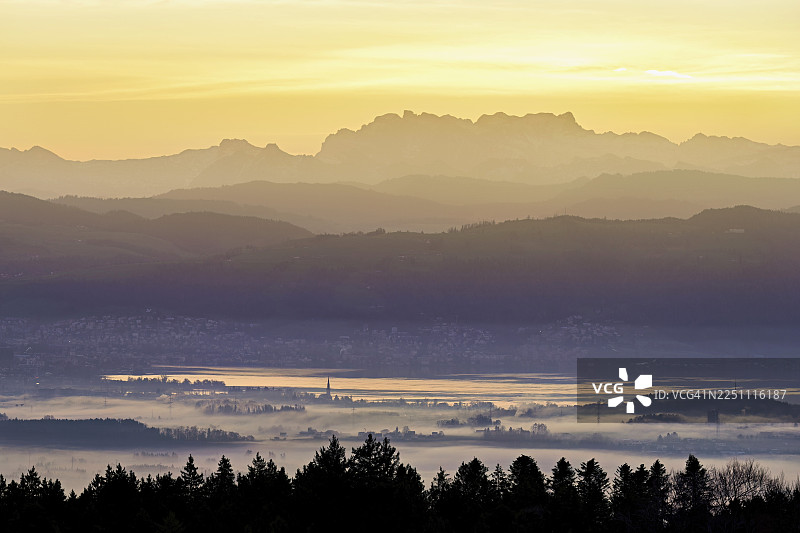 从霍尔本远眺楚格湖，楚格市和楚格市被大雾笼罩，其后是日出时分覆盖着白雪的弗吕布里格山和弗伦里斯加特利山，瑞士阿尔高州拜因维尔-弗莱阿姆特。图片素材