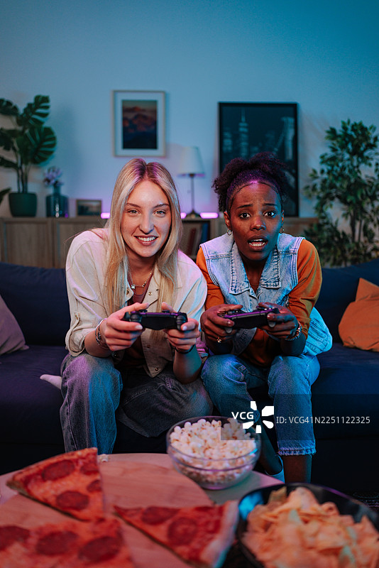 两个朋友在沙发上享受舒适的游戏之夜，旁边有零食和霓虹灯图片素材