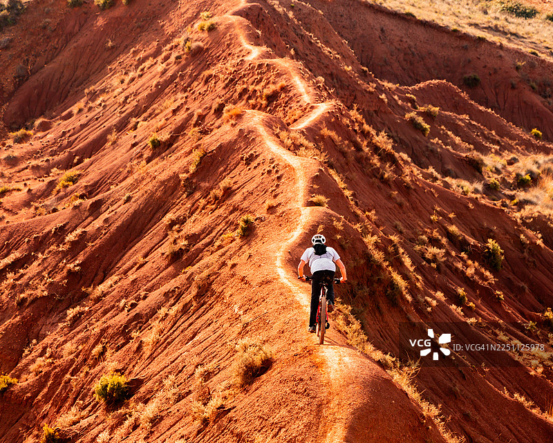 极限山地自行车手在偏远荒漠景观中穿越狭窄的红色山脊小径图片素材