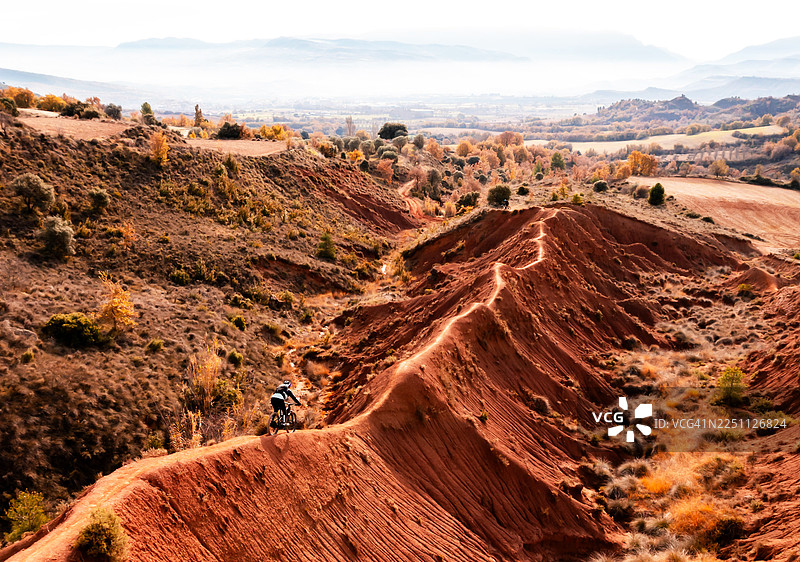 一名山地自行车手独自骑行在狭窄的红色山脊小径上，背景是壮观的红色岩石地貌。图片素材
