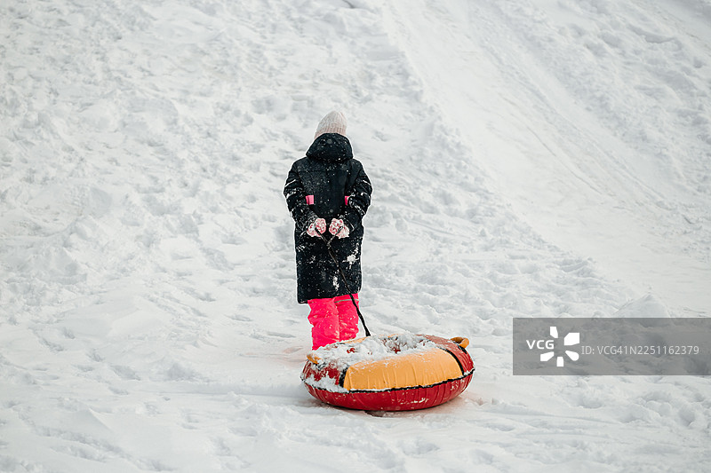 身穿冬季外套和粉色裤子的小女孩，在雪景中站着，旁边放着一个雪圈和一根拖绳图片素材