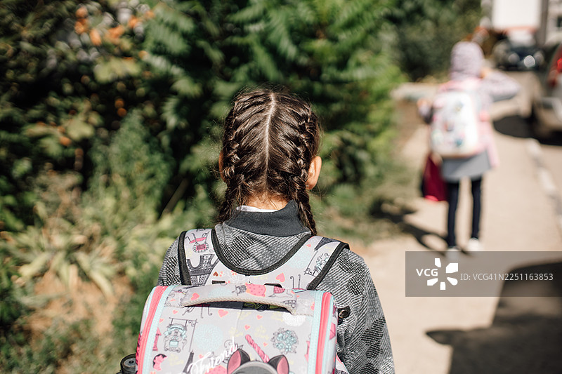 扎着辫子、背着彩色背包的女孩背影，走在阳光明媚的人行道上图片素材