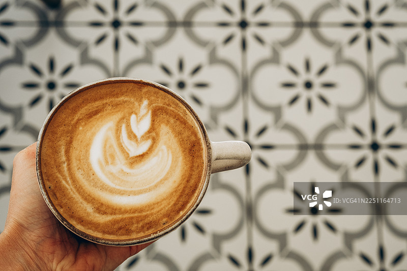 一只手拿着一杯卡布奇诺，上面有拿铁拉花，从上方拍摄，背景是装饰性图案的瓷砖地板，营造出舒适的咖啡馆氛围。图片素材