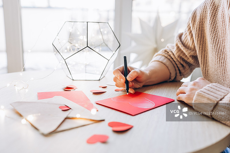 准备浪漫惊喜：特写一个人在木桌上用红色贺卡绘制心形，背景是纸心形纸屑、信封和柔和的串灯。图片素材