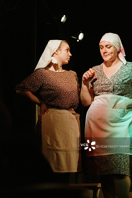 两位身着复古服饰的女性在戏剧舞台上，在温暖的灯光下交谈，定格戏剧性时刻图片素材