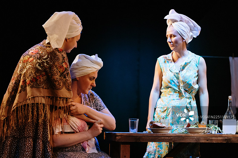 戏剧场景中，三位戴头巾的女性围坐在桌边，分享着片刻时光。图片素材