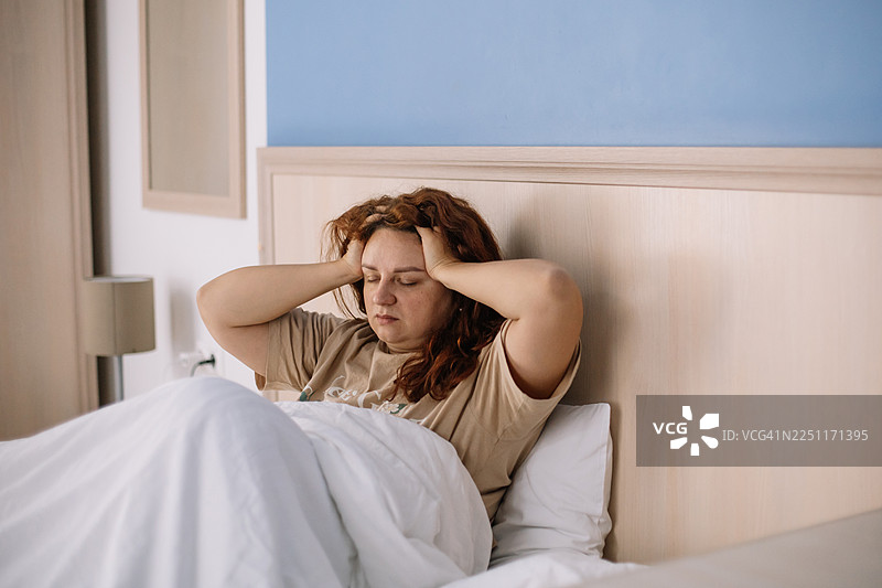 疲惫的女子躺在床上，捂着头，展现出疲劳、压力和宿醉的早晨感觉图片素材