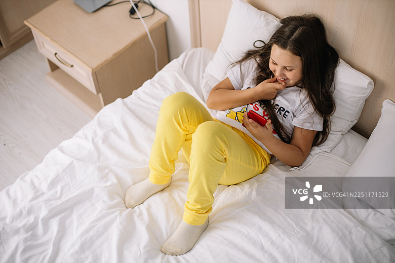 年轻女孩在舒适的卧室里躺在床上，对着手机微笑图片素材