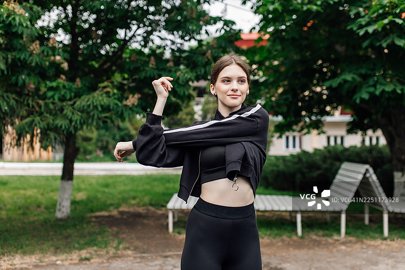 年轻女子身穿黑色运动服在公园里伸展手臂，进行轻度户外锻炼图片素材