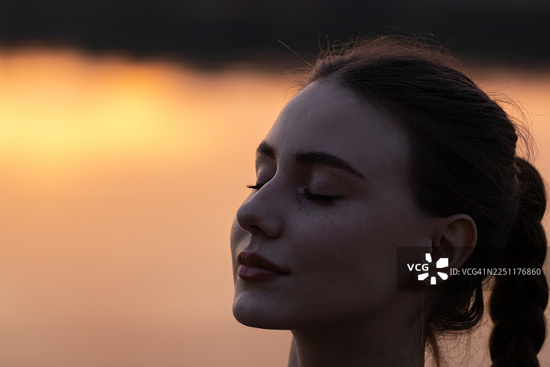 夕阳下，女子在水边闭目沉思的宁静肖像，传递出平静与温暖的光晕。图片素材
