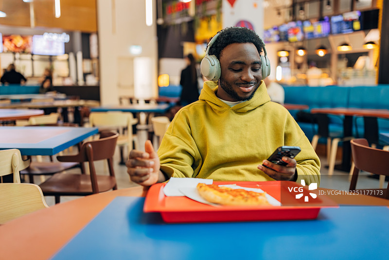 一位穿着黄色连帽衫的快乐黑人男子在热闹的商场美食广场午餐时吃着新鲜的披萨片图片素材