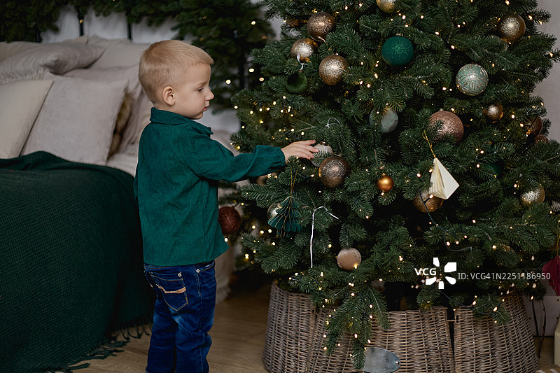 一个穿着深色衣服的金发男孩正在一个节日卧室里的圣诞树上触摸一个悬挂的装饰球。图片素材