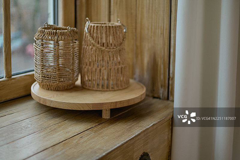 两个编织海草灯笼放在圆形木托盘上，托盘放在木制窗台上，旁边是白色窗帘。图片素材