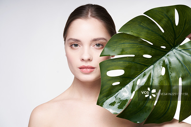 一位女性的特写工作室肖像，脸上部分遮挡着一片光泽的龟背竹叶，营造出神秘、自然的审美，适合美容和生活方式的影像。图片素材