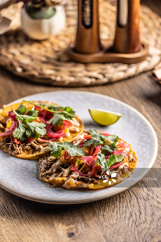 盘子里是两个酥脆的墨西哥玉米饼（玉米饼），上面铺满了肉馅、奶油酱、腌红洋葱和新鲜香菜。它们盛放在一个浅色、带斑点的盘子里，旁边还有一片青柠，背景是木桌。图片素材