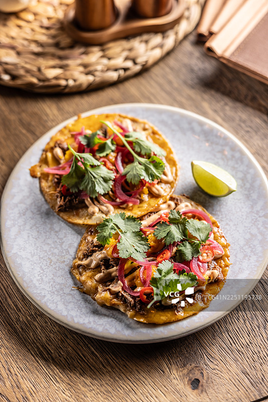 盘子里是两个酥脆的玉米饼（玉米饼），上面铺满了肉馅、奶油酱、腌红洋葱和新鲜香菜。它们盛放在一个浅色、有斑点的盘子里，配有一片青柠，背景是木桌。图片素材