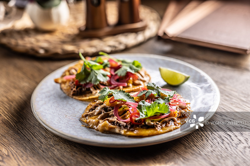 盘子里是两个酥脆的玉米饼（玉米饼），上面铺满了肉馅、奶油酱、腌红洋葱和新鲜香菜。它们盛放在一个浅色、有斑点的盘子里，配有青柠片，背景是木桌。图片素材
