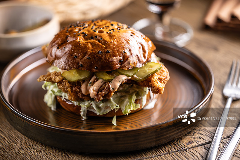 这份鸡肉汉堡采用光泽感的黑芝麻面包和炸鸡，搭配酸黄瓜和卷心菜丝，带来清爽的口感。它被优雅地摆放在一个带有纹理的深色盘子上，背景中还可以看到一杯拉格啤酒。图片素材