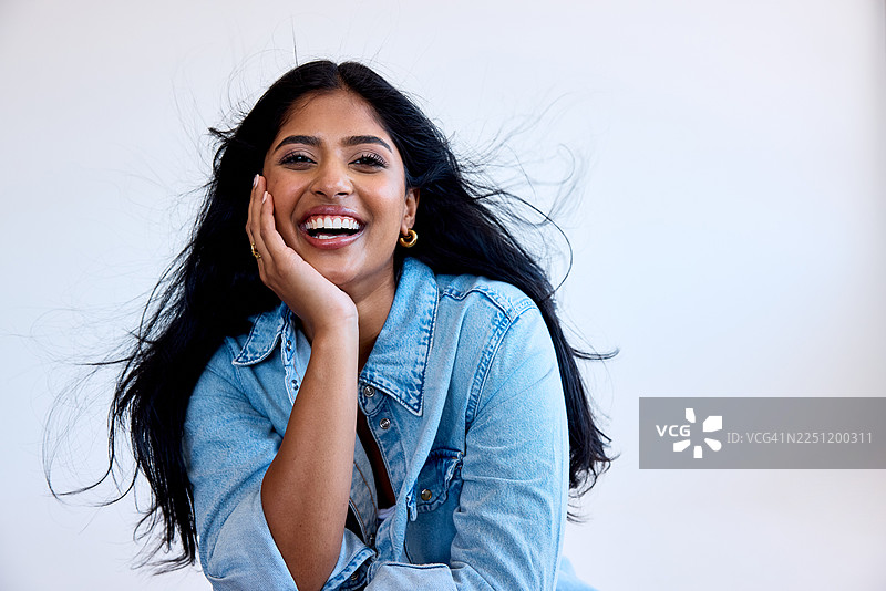 身穿牛仔夹克的年轻女子，秀发飘逸，单手托腮，脸上洋溢着喜悦，摄于工作室图片素材