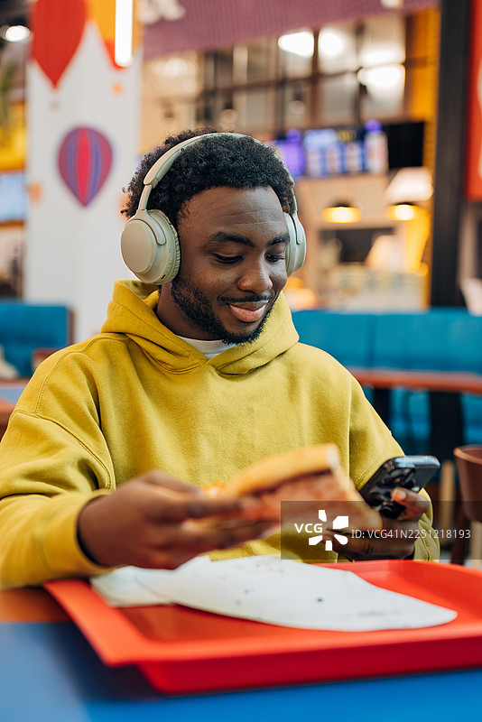 一位英俊的黑人男性在现代美食广场享用休闲快餐午餐，同时使用手机应用和听音乐。图片素材