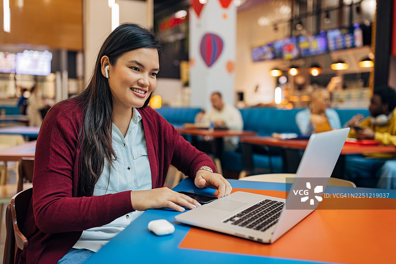 一位亚洲女商人坐在明亮美食广场的彩色桌子旁，用笔记本电脑工作，代表着现代远程办公或自由职业者的生活方式。图片素材