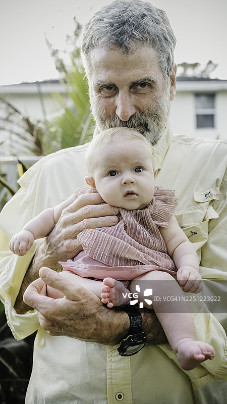 一位六十多岁的银发祖父在户外拍摄肖像，怀里抱着他可爱的小孙女。图片素材