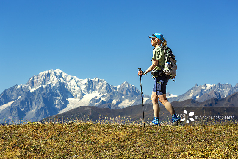 一位徒步者手持登山杖，走在长满青草的山丘上，背景是白雪皑皑的山脉。地点：瓦莱达奥斯塔，意大利。图片素材
