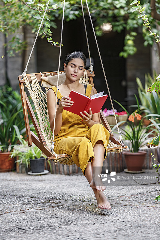 身穿黄色连衣裙的女子坐在花园秋千上阅读一本红色的书，周围摆放着盆栽植物。地点：西班牙阿尔梅里亚，塞拉尔哈米利亚。图片素材