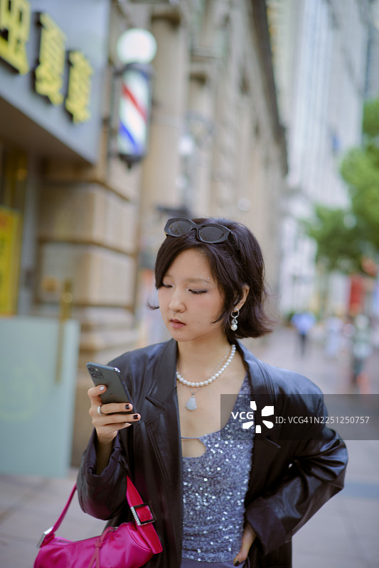 年轻女子在城市环境中查看手机，身穿灰色上衣、黑色夹克和粉色包。中国上海。图片素材