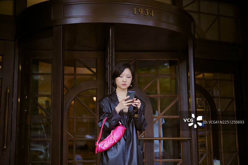 穿着黑色外套的女士在旋转门前使用智能手机，她挎着一个亮粉色的包。中国上海。图片素材
