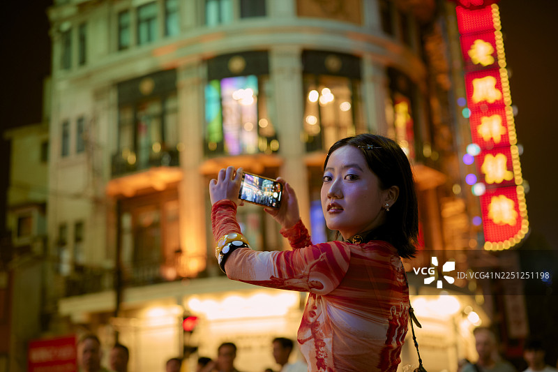 一位女士在中国上海充满霓虹灯招牌和高大建筑的活力街道上拍摄夜景照片。图片素材