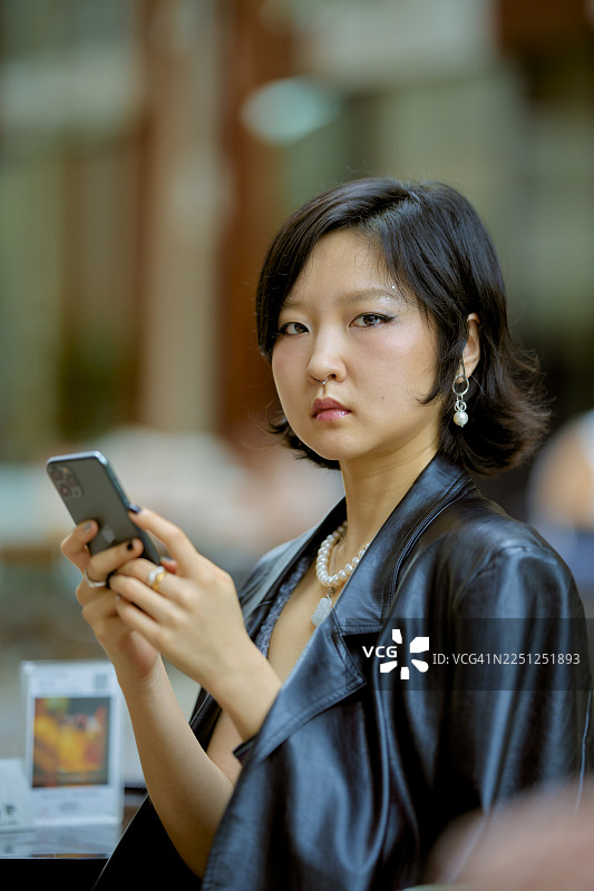 身穿黑色夹克的年轻女子，手持智能手机，看向镜头。中国上海图片素材