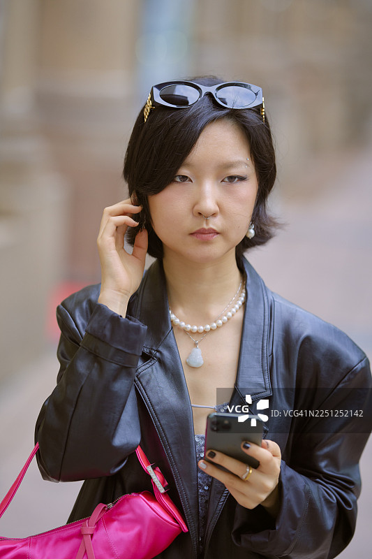 穿着皮夹克的年轻女子，头戴太阳镜，手持手机和粉色包，摄于中国上海图片素材
