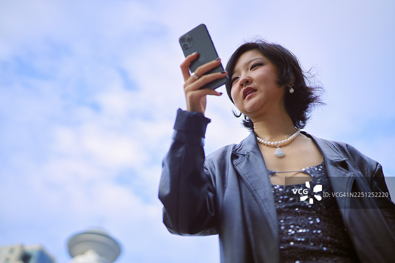 身着时尚服装的女性在蓝色天空背景下看着智能手机。中国上海。图片素材