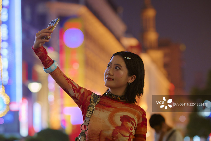 夜间，一位年轻女子在中国上海灯火辉煌的城市街道上自拍。图片素材