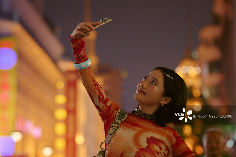 身着鲜艳服装的年轻女子在夜晚的上海街头，被灯光环绕，正在自拍。图片素材