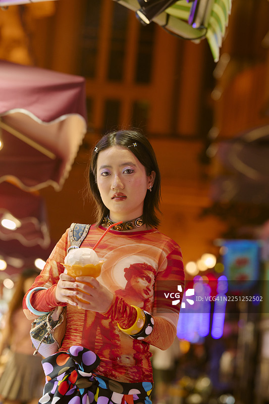 年轻女子手持饮品，站在上海夜市的彩色雨伞下。图片素材