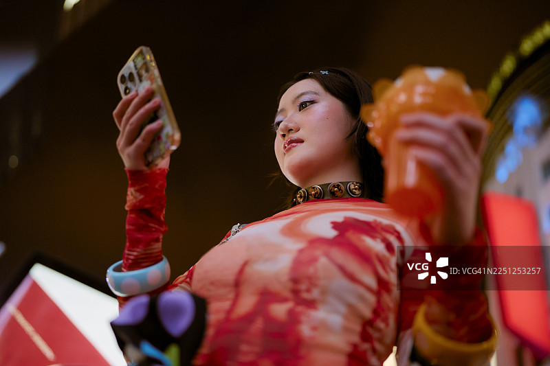 身着鲜艳服饰的年轻女子在城市背景下，一边拿着手机和饮品，一边抬头望去。中国上海。图片素材