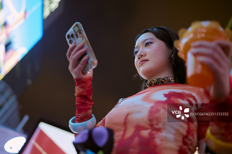 夜晚的城市里，一位穿着鲜艳服装的年轻女子手持饮品和智能手机。中国，上海。图片素材