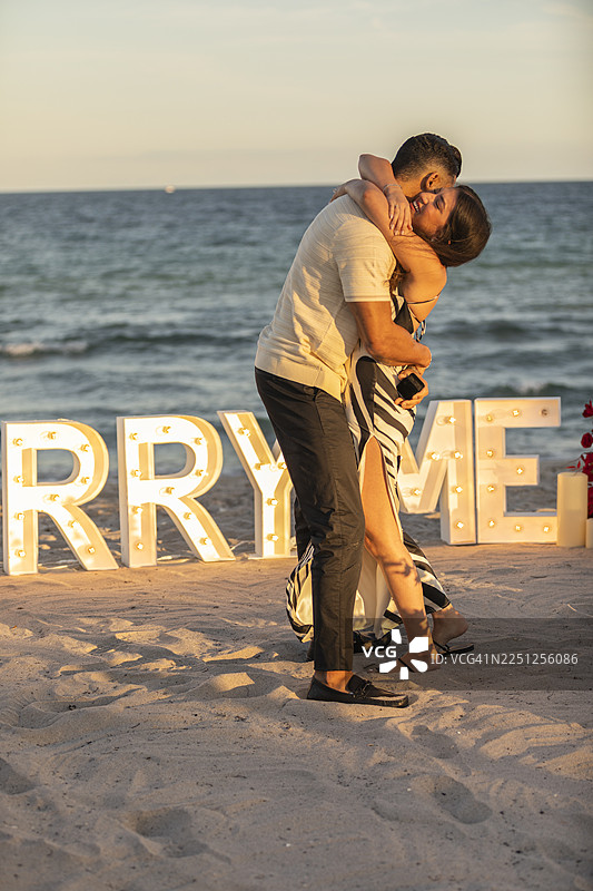 一对新订婚的情侣在日落时分的海滩上喜悦地拥抱，周围环绕着红色的花朵和闪耀的“Marry Me”字母。图片素材