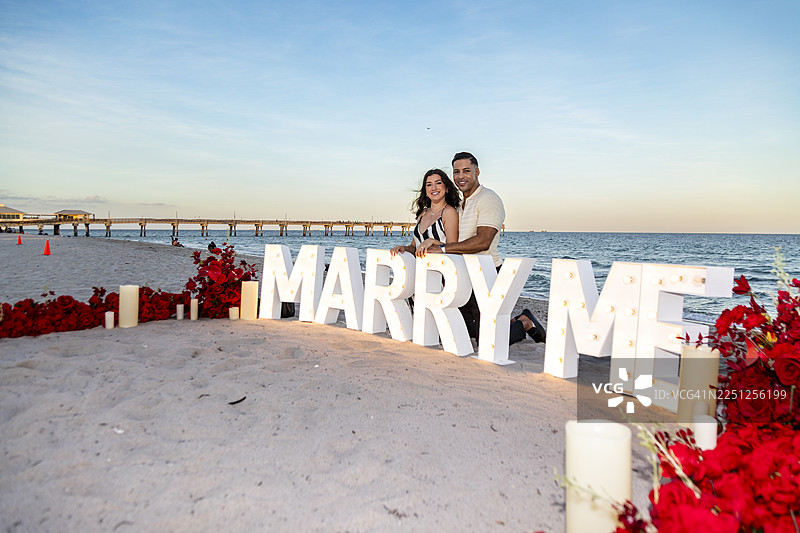 一对新订婚的夫妇在日落时分，在发光的“嫁给我”字母前，开心地在海滩上合影。图片素材