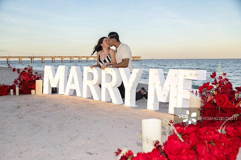 一对新订婚的情侣在日落时分的海滩上，在发光的“Marry Me”字母前浪漫拥吻。图片素材