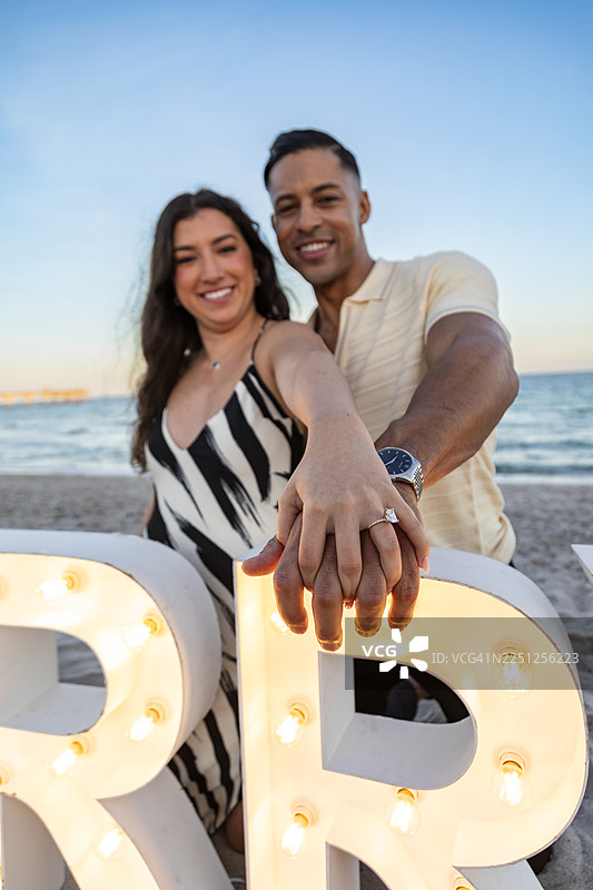 一对新订婚的情侣在海滩上深情地笑着，他们手牵着手，展示着订婚戒指。图片素材